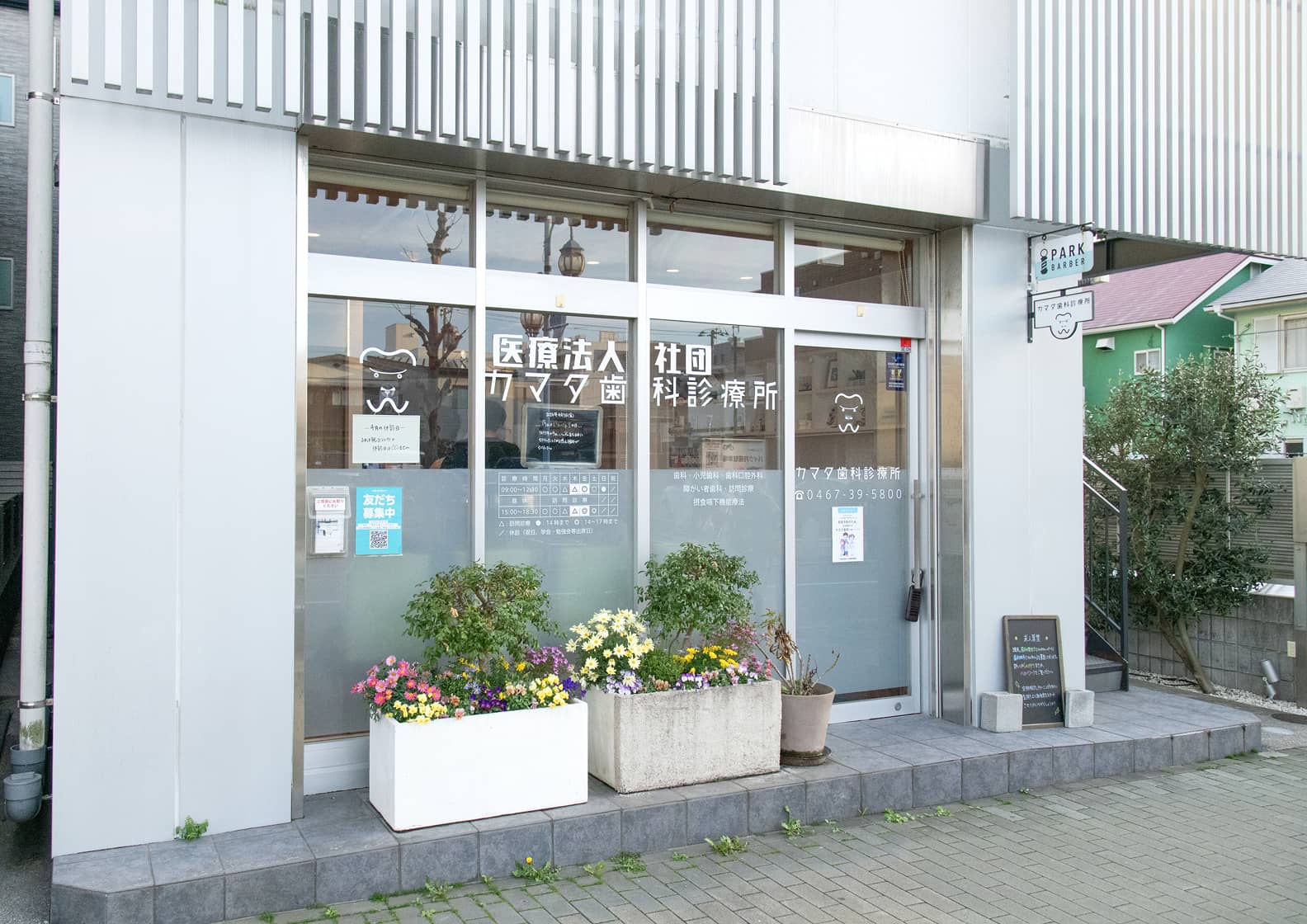 医療法人社団カマタ歯科診療所の外観。白い外壁の清潔感のある建物で、入り口のガラス戸には診療時間やロゴが掲示されています。足元には色鮮やかな花が植えられたプランターが並び、明るく入りやすい雰囲気の入り口です。