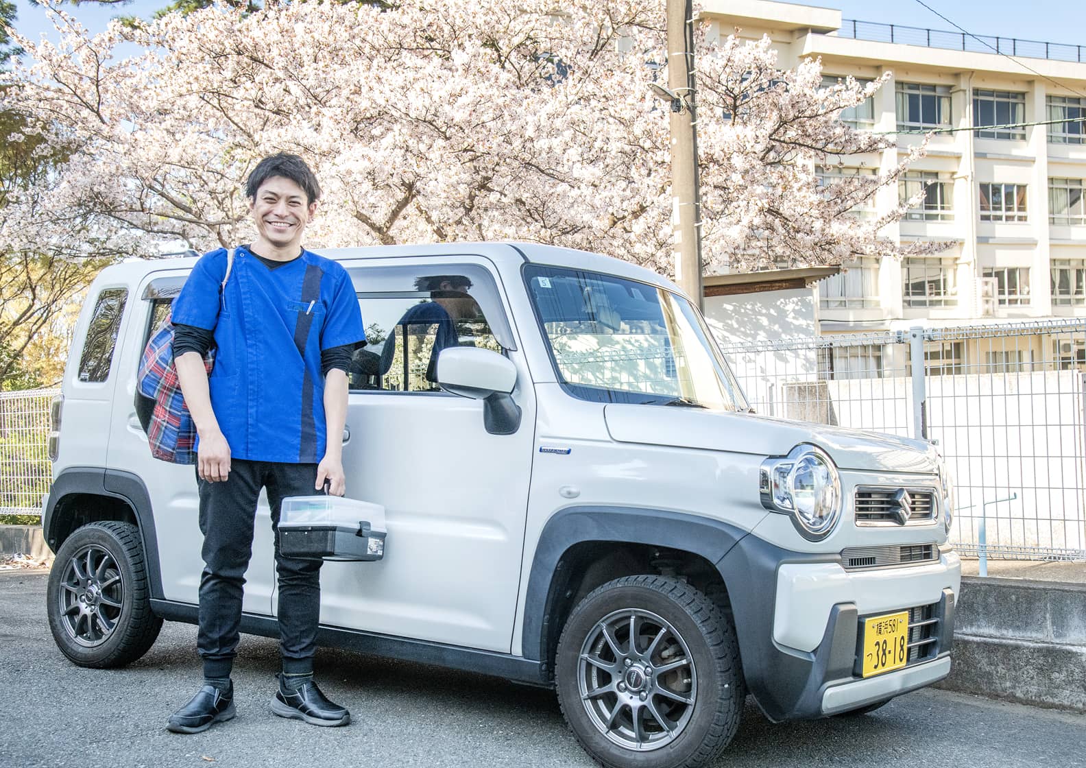 満開の桜を背景に、訪問診療車（軽自動車）の前に立つ歯科医師。青いスクラブを着用し、手に往診用機材ケースを持って笑顔を見せています。地域に根ざした訪問歯科診療への意気込みが伝わる一枚です。