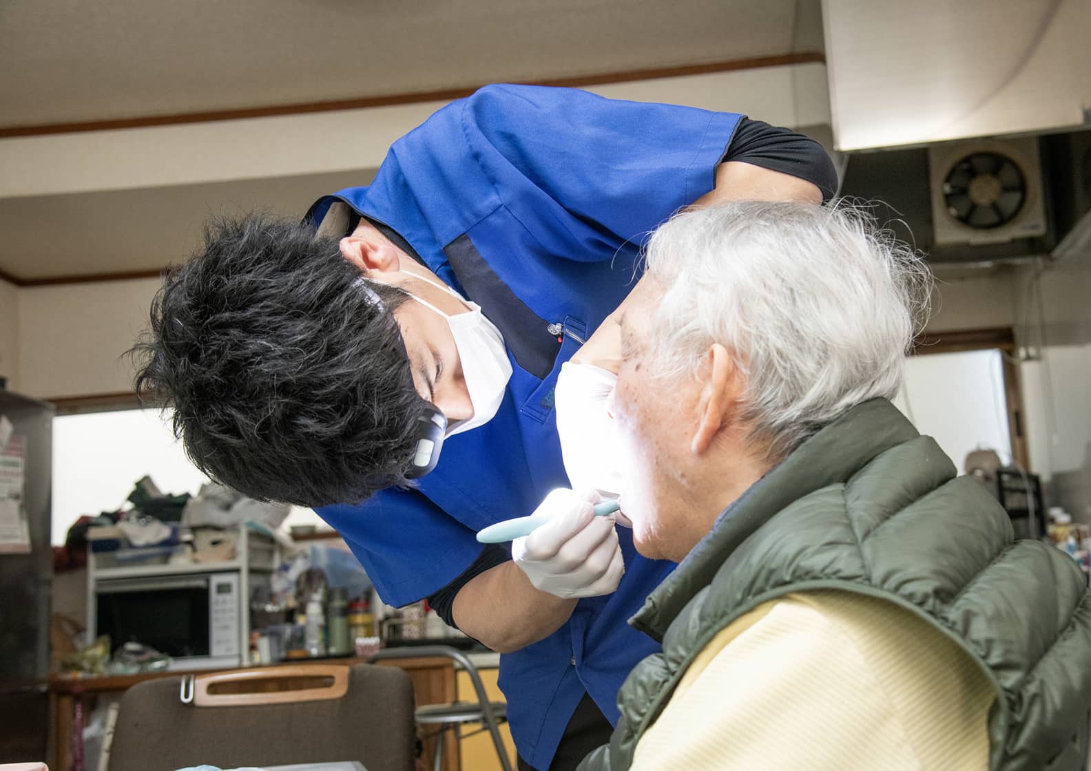 歯科医師による訪問診療の様子。青いスクラブを着用した医師が、自宅で療養中の高齢男性の口内を丁寧に診察しています。手元をライトで照らし、患者の表情を確認しながら優しく声をかけているような安心感のあるシーンです。