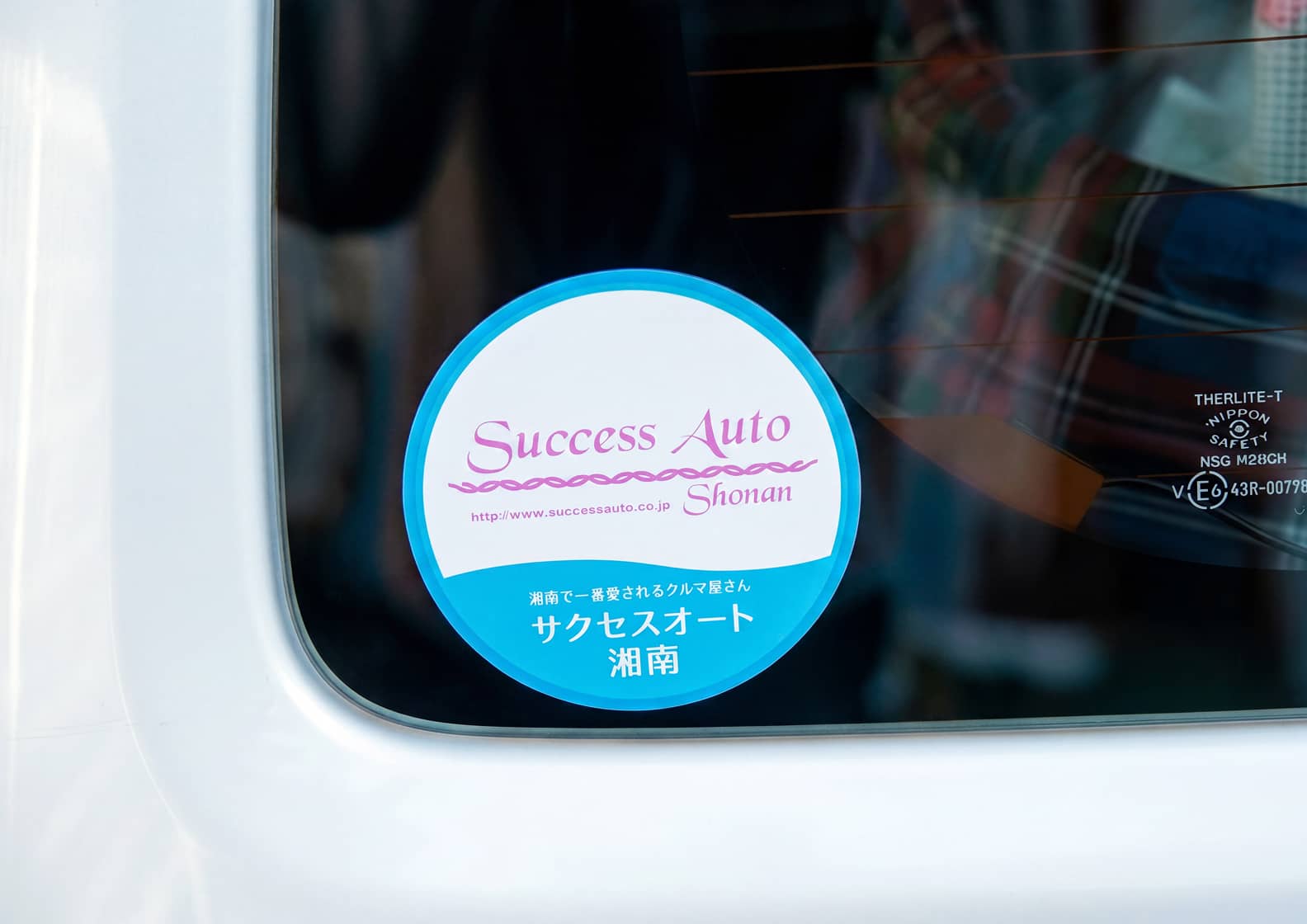 車両の窓ガラスに貼られた丸いステッカー。「Success Auto Shonan（サクセスオート湘南）」と書かれ、ピンクと水色の配色で「湘南で一番愛されるクルマ屋さん」というメッセージが記されています。