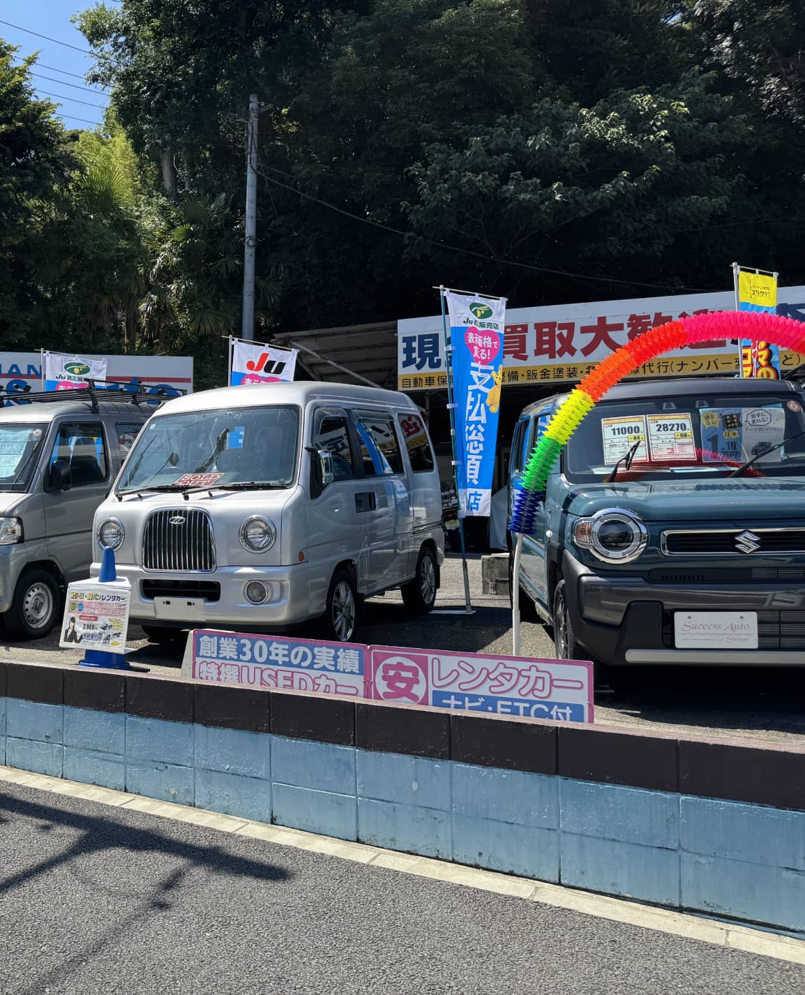 中古車販売店に並ぶシルバーの軽バンとグリーンのスズキ車の展示風景